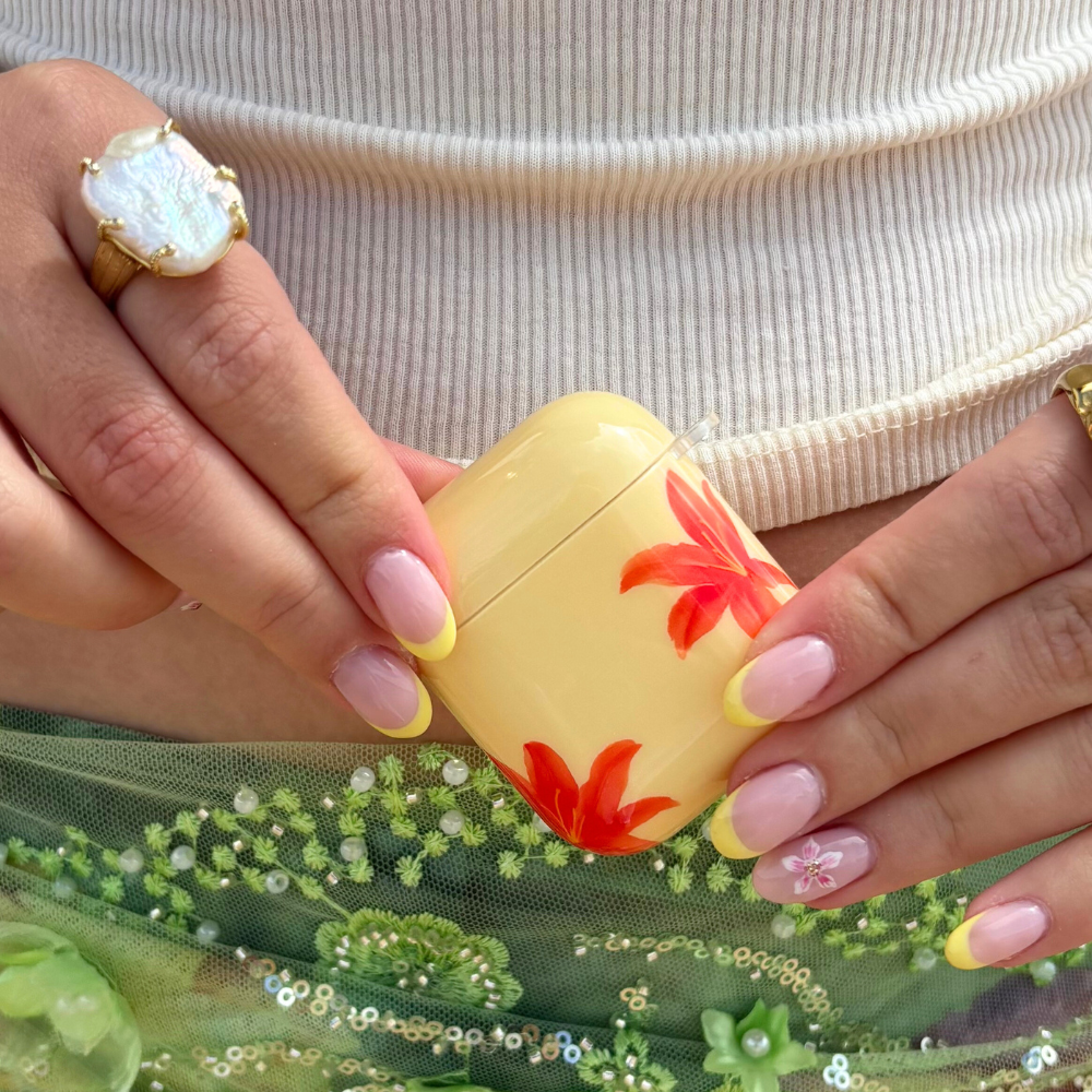 Hands with pink and yellow nails hold a floral Coconut Lane AirPods case, Island Girl Ibiza Lily, next to a ring.