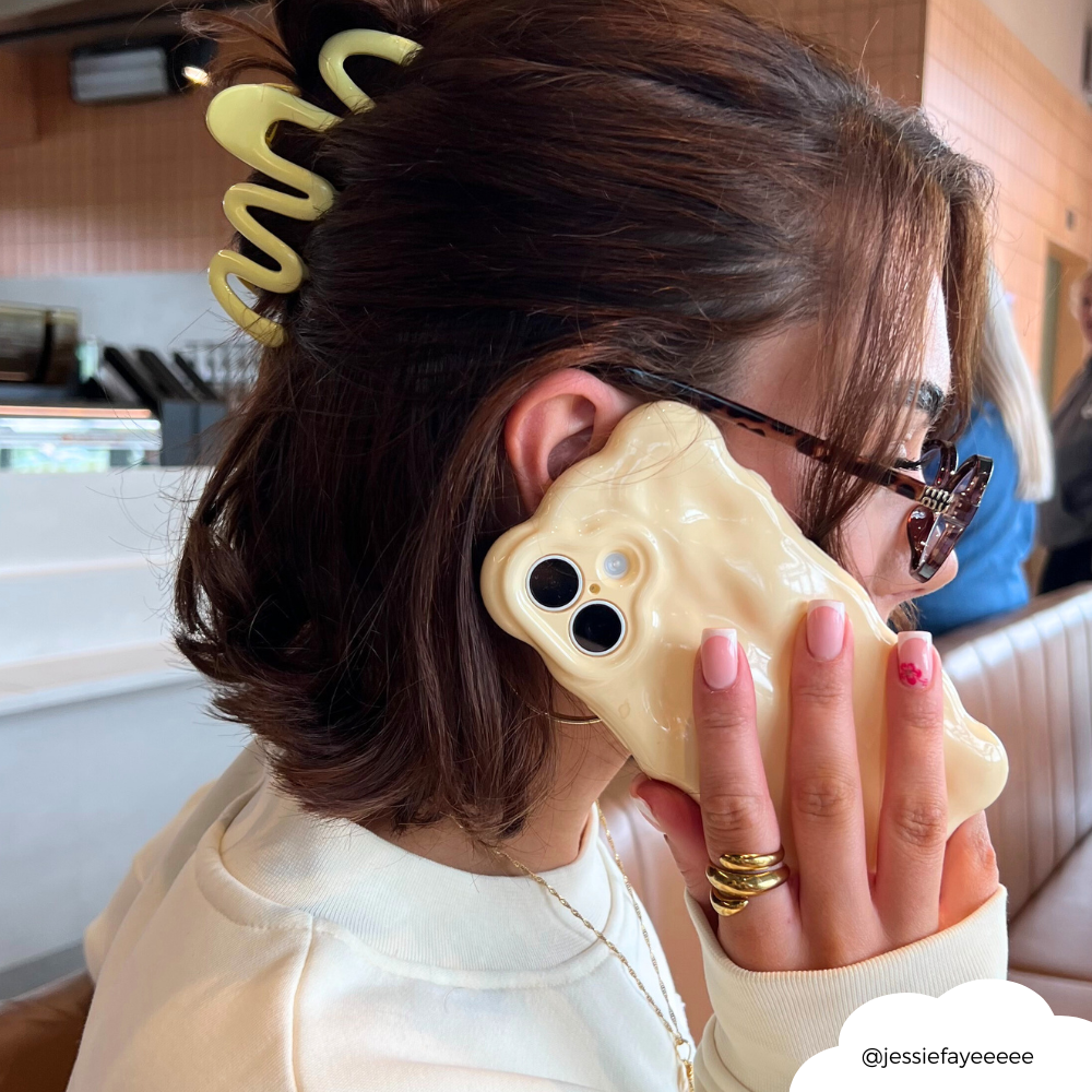 Woman with short brown hair wears a yellow squiggle claw clip and holds a wavy cream phone case.