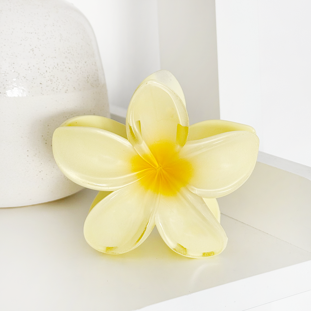 Butter yellow flower-shaped claw clip on a white shelf beside a white ceramic vase.