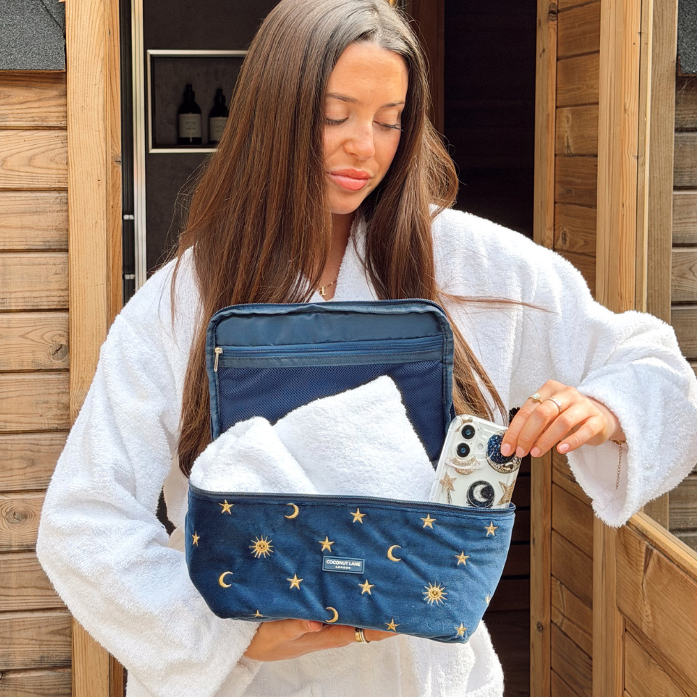 girl outside sauna in white dressing gown holding celestial vanity case with towels inside and celestial phone case next to it