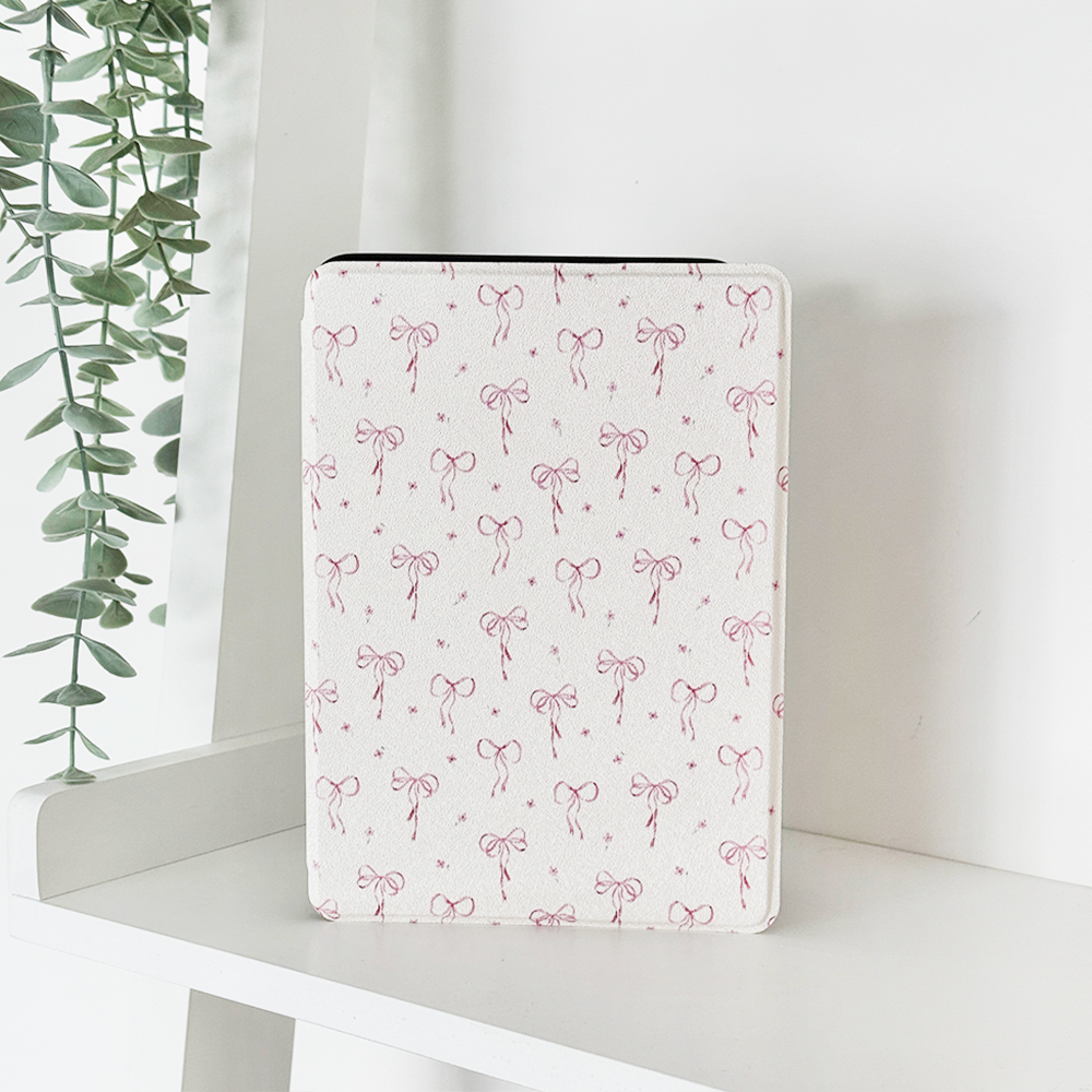 A white tablet case with a pink bow pattern stands upright on a white shelf, with a green trailing plant in the background.