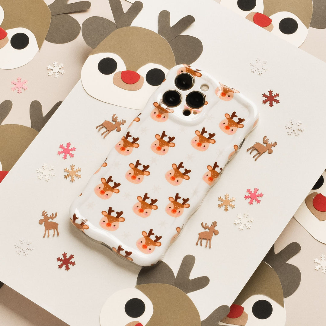 A phone with a clear case decorated with cute reindeer faces lies on festive paper featuring reindeer faces, snowflakes, and small reindeer illustrations in a holiday theme.