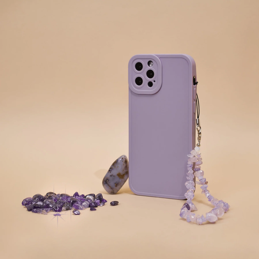 A lavender smartphone case with a wrist strap adorned with purple gems is pictured against a beige background. Nearby, a small pile of purple gemstones and a large stone are arranged artistically.