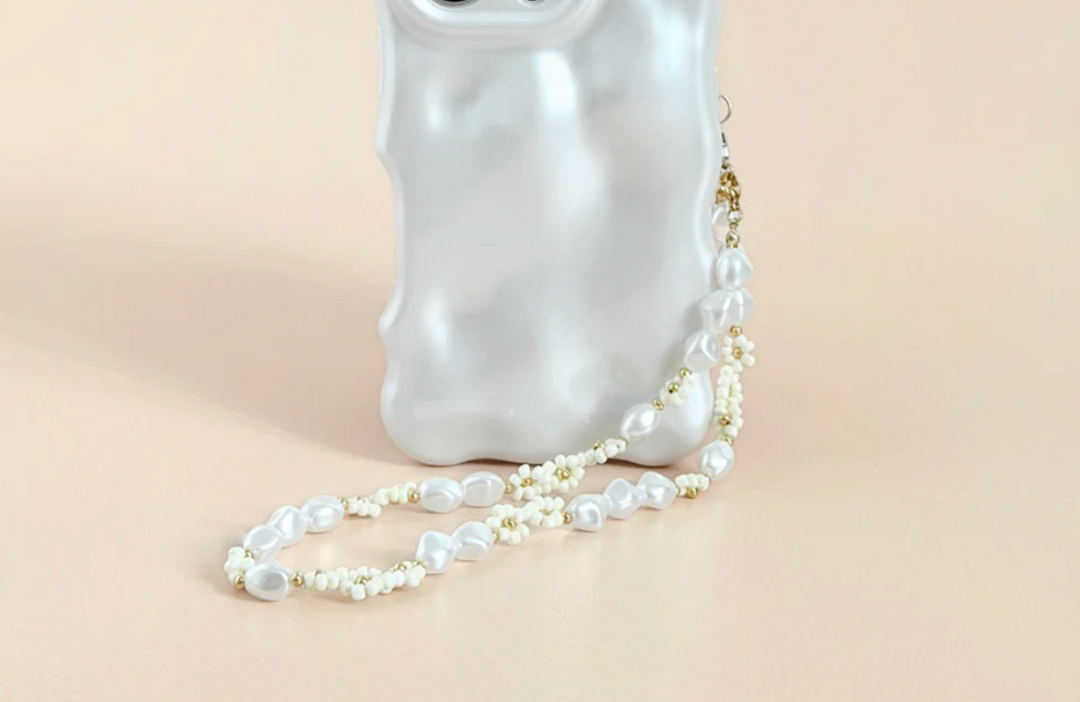 A hand with manicured nails holds a beige smartphone with a decorative case and a translucent beaded charm. The background shows a patterned fabric, possibly part of the person's clothing.