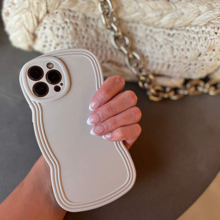 A hand with manicured nails holds a white smartphone encased in the Coconut Lane NAKD Curvy Phone Case - Vanilla, celebrated for its high protection and lightweight design. In the background, a textured beige bag adorned with a chunky gold chain strap adds an elegant touch.