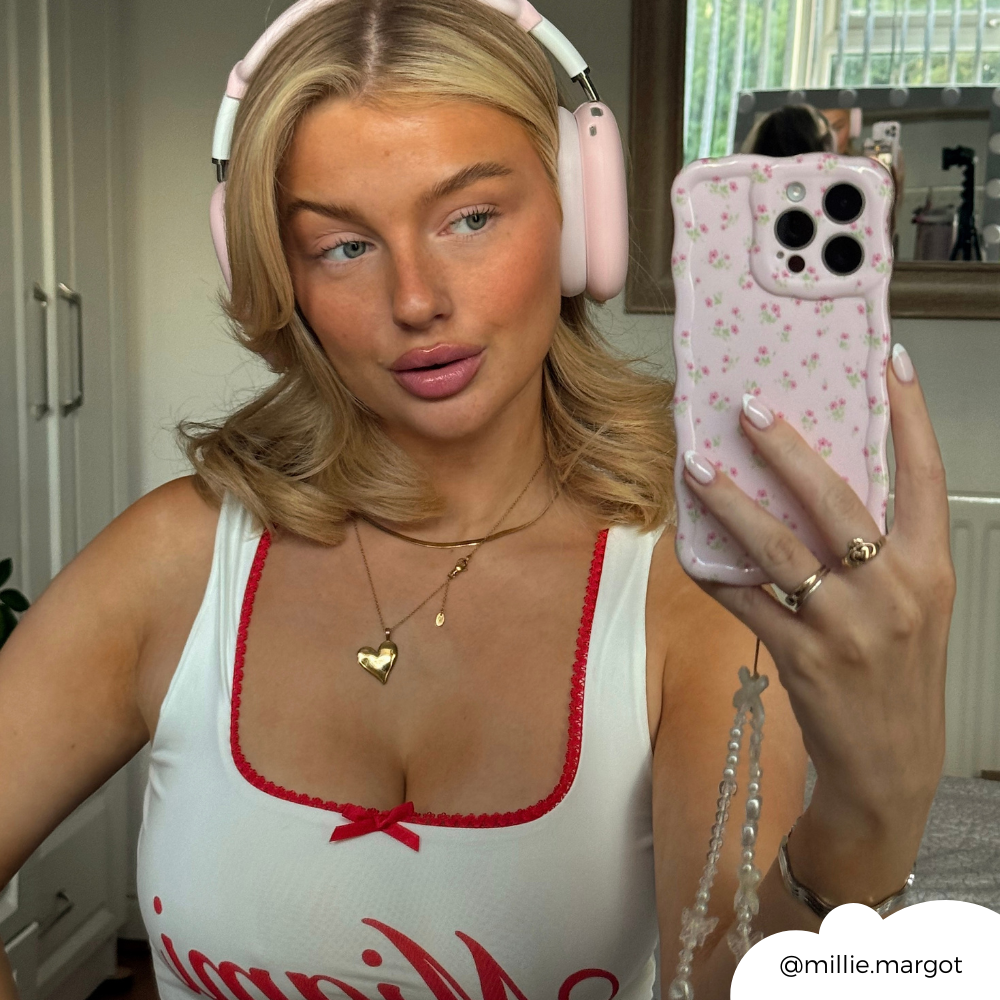 Blonde woman in headphones holds pink floral wavy phone case, taking a mirror selfie.