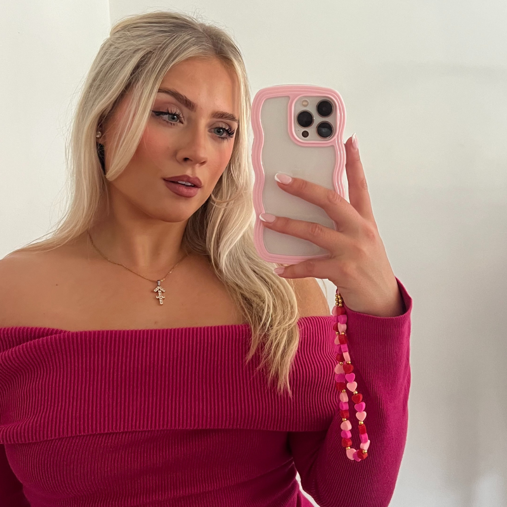 A woman with long blonde hair takes a mirror selfie with her phone in a pink case that features the Coconut Lane Beaded Phone Strap - Hearts. She is dressed in an off-the-shoulder, ribbed, long-sleeve top in a deep magenta color and accessorizes with a golden necklace adorned with a cross pendant. The background is a plain white wall.