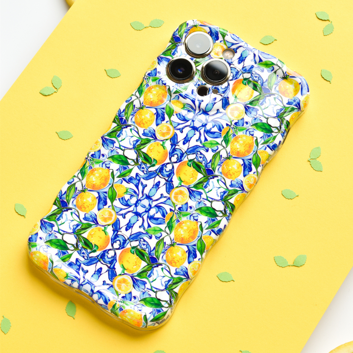 A smartphone adorned with Coconut Lane's Wavy Phone Case - Amalfi Lemon, boasting a vibrant pattern of yellow Amalfi lemons and blue flowers, is placed on a yellow surface. Green leaf-shaped confetti is scattered around the phone.