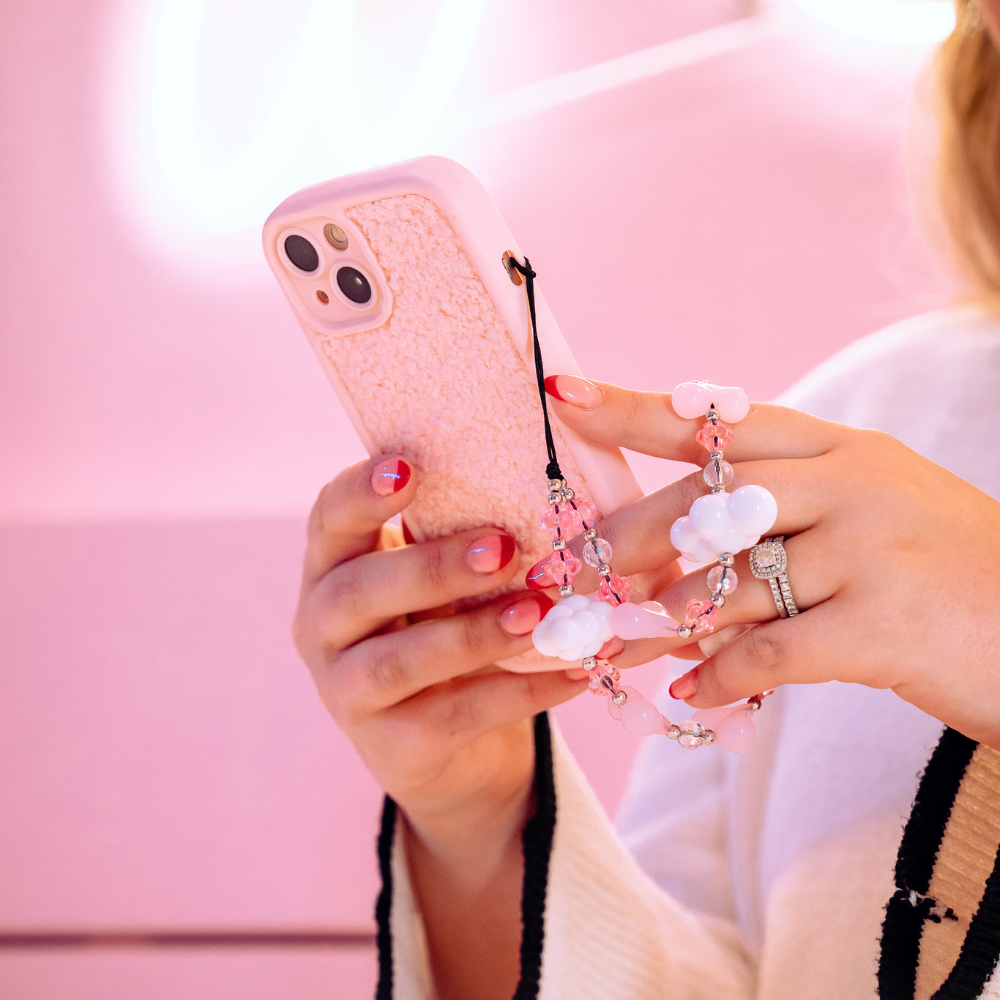 Close-up of a person holding a Coconut Lane Cosy Teddy Phone Case - Pink, adorned with a beaded charm. The person has manicured nails painted in pink and wears a ring with a large gemstone. The background features soft pastel pink tones.
