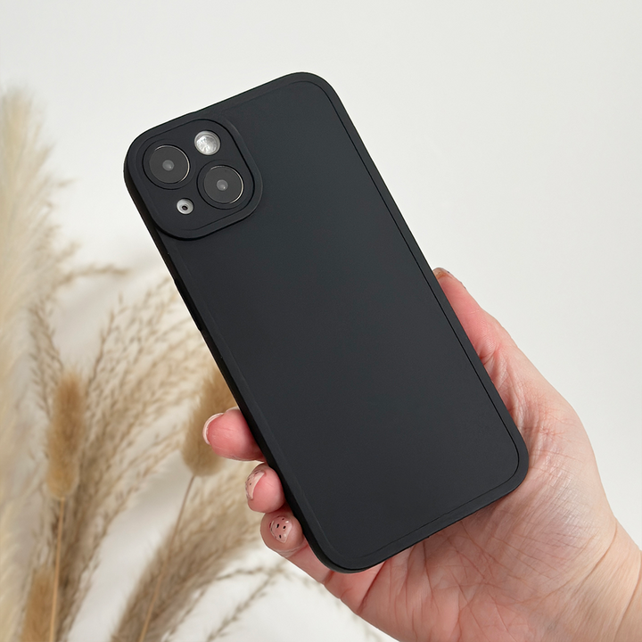 A hand holding a black smartphone with a dual-camera setup, encased in a NAKD Phone Case - Midnight Black from Coconut Lane, made of Soft Flexible TPU, slightly angled to display the back. The background features a neutral-colored setting with some pampas grass, adding a natural, minimalist touch to the scene.