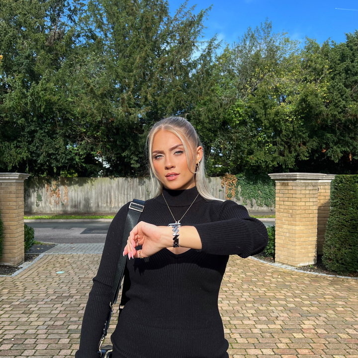 A person with long blonde hair, wearing a black turtleneck sweater and a Coconut Lane Get Wavy Apple Watch Strap in black, stands on a cobblestone driveway, looking at their watch. Tall green trees and brick pillars are in the background under a blue sky.