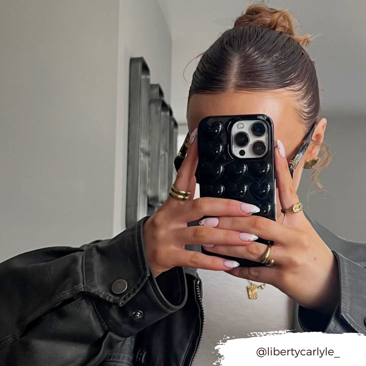 A person with light pink nail polish holds an iPhone with a Coconut Lane Glossy Black 3D Hearts Phone Case, blocking their face. They wear a dark jacket over a white top, and their hair is styled neatly. The handle @libertycarlyle_ is visible in the bottom right corner.