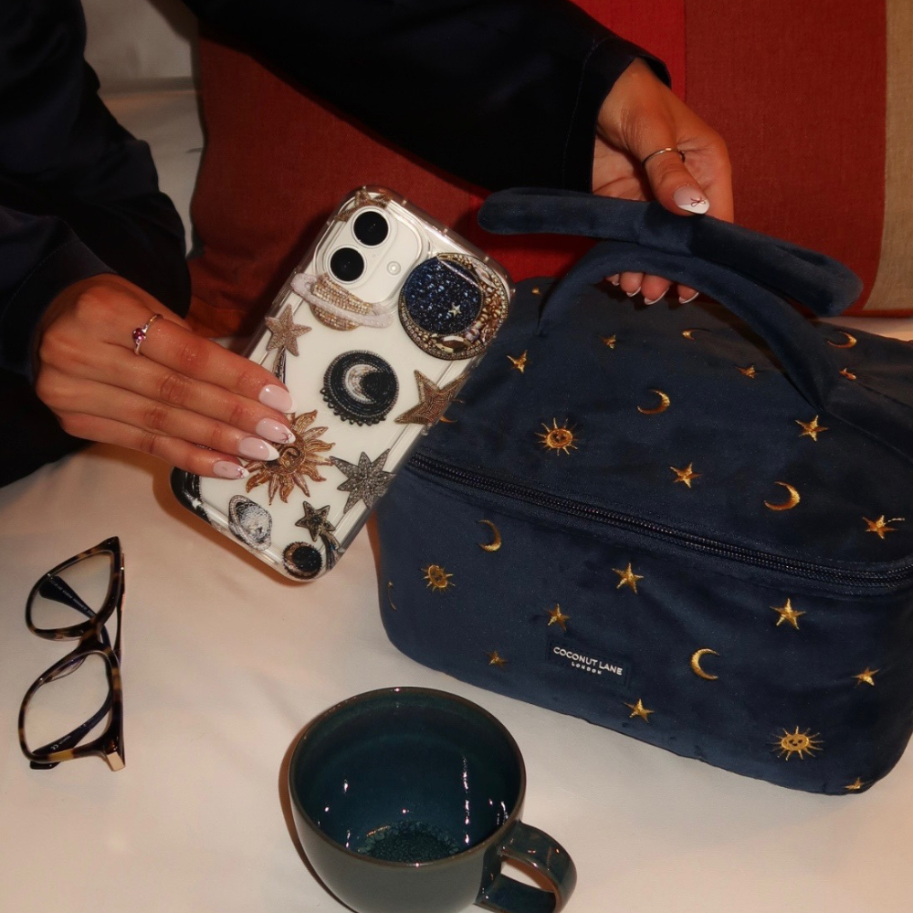 Clear phone case with celestial design held next to a navy moon-and-stars bag, glasses, and green cup.
