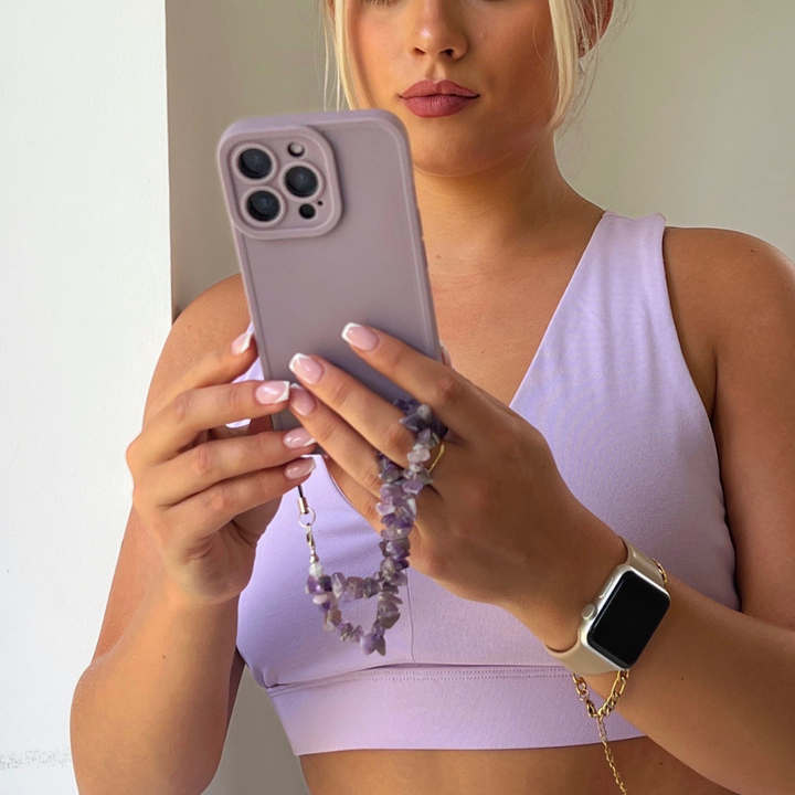A woman wearing a lavender sports bra and an Apple Watch is holding a lavender-colored iPhone adorned with a Coconut Lane Crystal Phone Strap in Lilac. She is standing against a white background and looking at her phone screen, showcasing her decorative accessory.