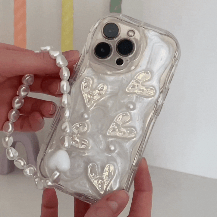A person is holding the Pearly Hearts Phone Case - Clear by Coconut Lane, which features a pearlescent finish embellished with gold heart designs. This extra-protective iPhone 15 case also includes a detachable beaded strap resembling pearls. The background is a colorful blur of pastel hues.