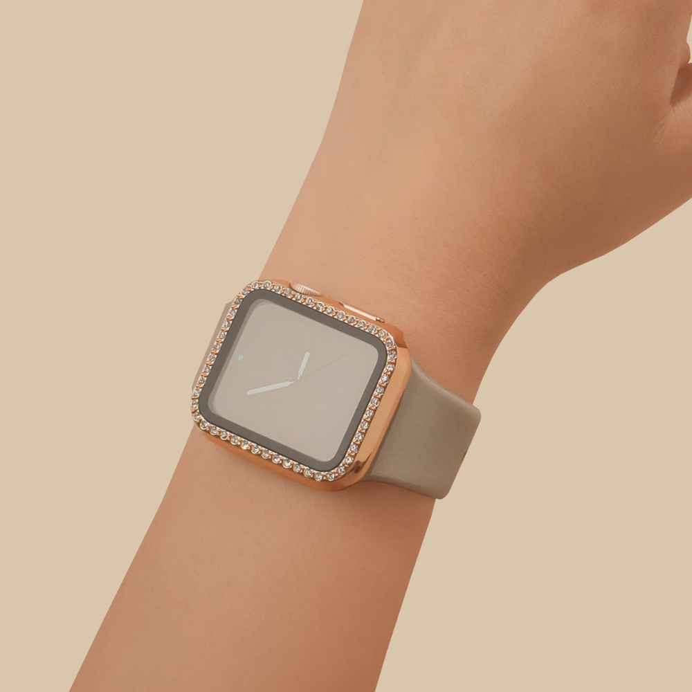 A close-up of a wrist wearing the Coconut Lane 2-in-1 Apple Watch Case & Built-In Screen Protector - Rose Gold Diamante, featuring a rectangular face. The watch case has a rose gold diamante frame adorned with small, sparkling stones around the edges and is paired with a light gray strap. The background is a solid beige color.