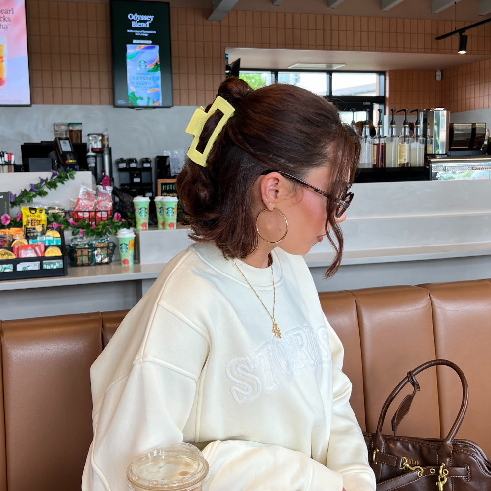 Woman in a cafe booth wears a yellow square claw clip, with an iced drink and brown bag on the table.