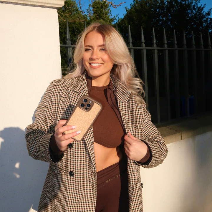 A smiling person with long, blond hair holds a phone adorned with the Coconut Lane Cosy Teddy Phone Case in Salted Caramel in one hand and adjusts their brown checked coat with the other. They are wearing a matching brown crop top and pants, standing in bright sunlight in front of a white wall and dark fence.