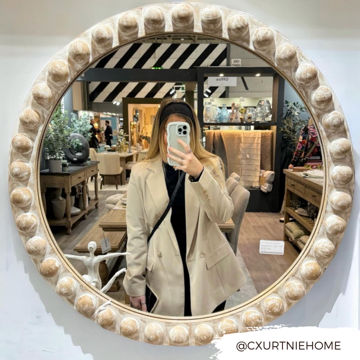 A person wearing a beige blazer and black top takes a mirror selfie in a home decor store. Various items like furniture and decorative pieces are displayed in the background. The mirror has a round, ornate frame with a beaded design. They're holding the Curvy Phone Case - Cream by Coconut Lane, which is highly protective for their iPhone 15. Text in the corner reads "@CXURTNIEHOME.