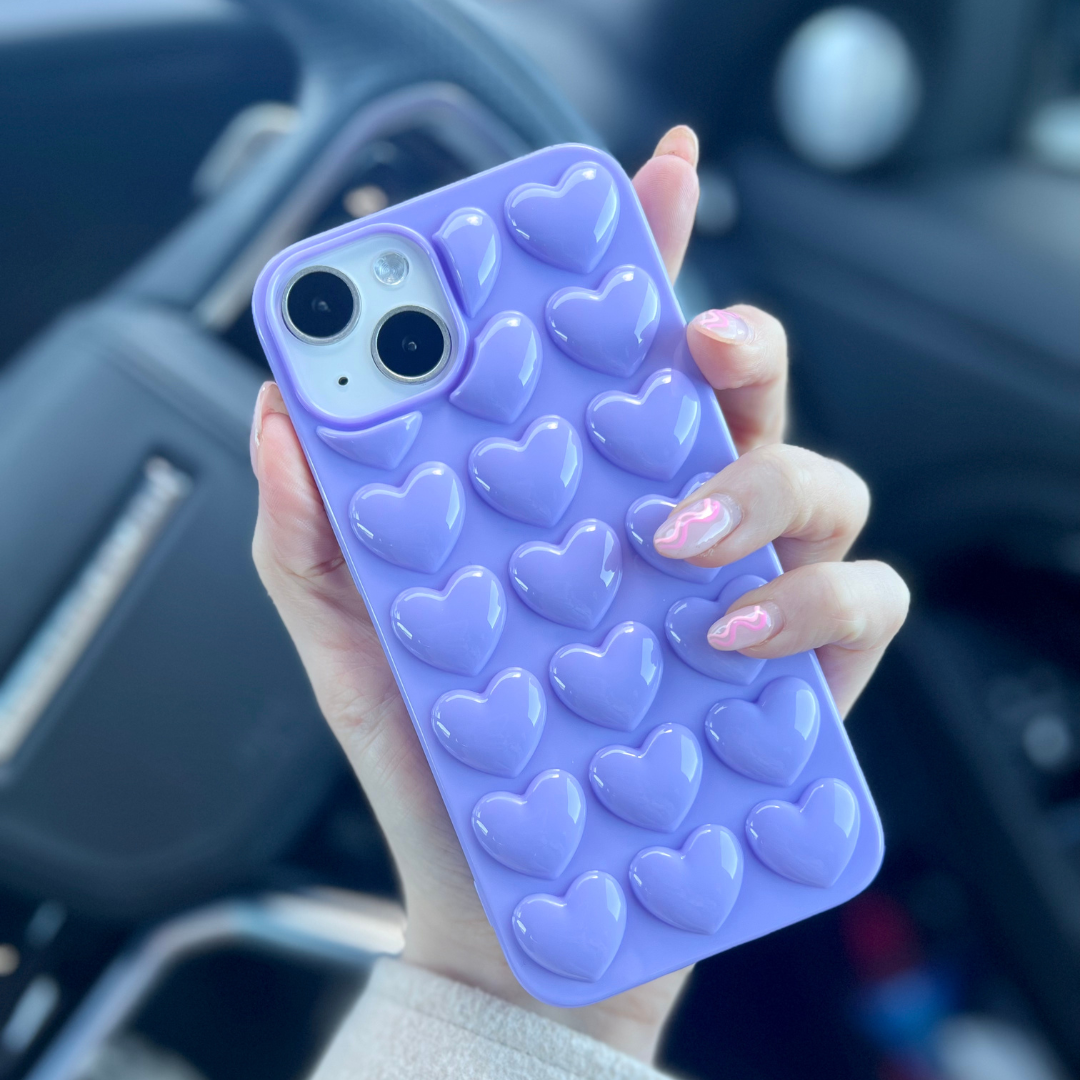 A hand with long, light pink manicured nails holds a Coconut Lane 3D Hearts Phone Case in Deep Lilac, adorned with raised heart-shaped designs. In the background, a blurred car interior featuring a steering wheel and controls can be seen.