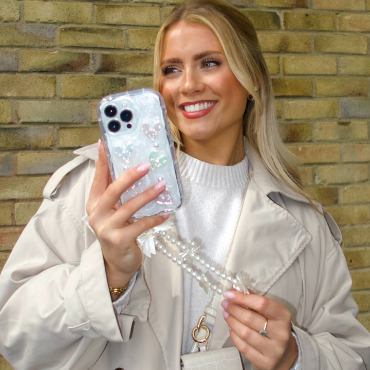 A blonde woman with a bright smile holds an iPhone 15 adorned with Coconut Lane's clear Pearly Hearts Phone Case and a pearl chain. She is leaning against a brick wall, wearing a light beige trench coat over a white sweater, accessorized with a small white handbag and a ring on her finger.