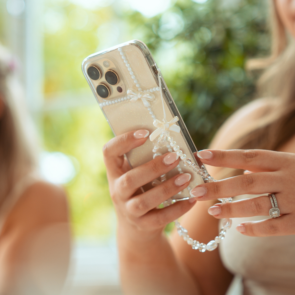 Close-up of a woman's hands holding an iPhone encased in a clear, beaded case adorned with small bows. The woman, who is wearing an engagement ring and Coconut Lane's Beaded Phone Strap - Bows & Butterflies, has manicured nails. The background features a soft blur of greenery and another person partially visible.