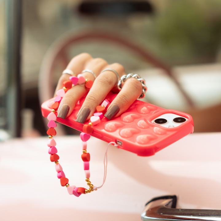 A hand with dark painted nails holding a red phone case adorned with a heart pattern and a beaded pink and red heart charm. The hand, wearing several rings, rests on a white and pink surface with a blurred background, showcasing the elegant Beaded Phone Strap - Hearts by Coconut Lane.