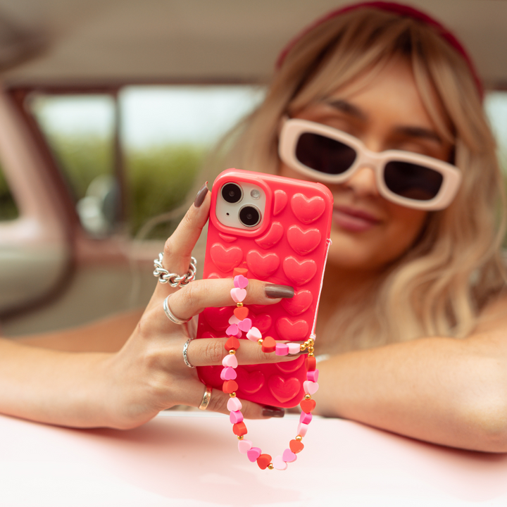 A person with long, blonde hair wearing a red headband and white sunglasses is sitting in a car, holding a phone adorned with a red heart-patterned case and the Beaded Phone Strap - Hearts from Coconut Lane. They have painted nails and are accessorized with silver rings and a bracelet.