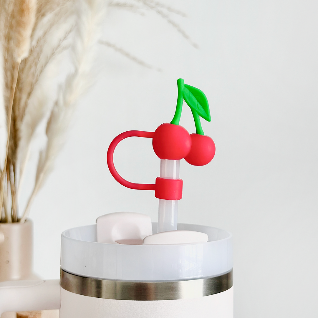 The Cherries Straw Cover by Coconut Lane features a white tumbler adorned with a silver rim, complemented by a uniquely designed silicone straw topped with a red cherry-shaped decoration and green leaves for added durability. The cover ensures the straw remains hygienic, while in the background, a vase filled with dried pampas grass enhances the minimalist and clean aesthetic.