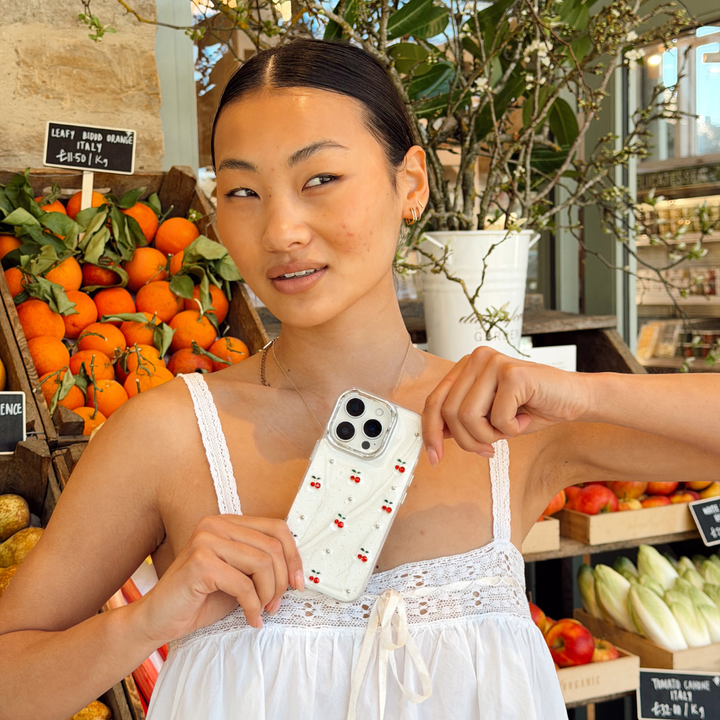 Girl next to a fruit stand holding the Cherry Pearl Phone Case posing
