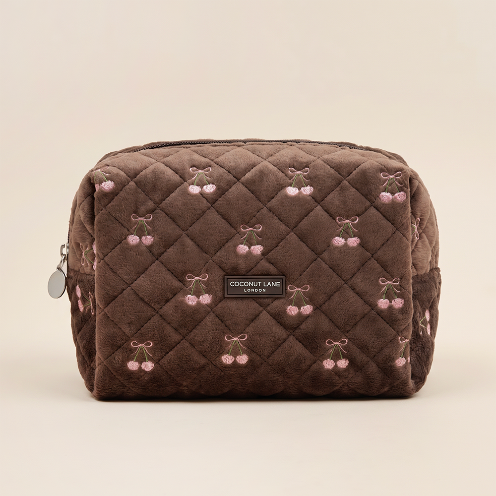 Brown quilted velvet makeup bag with pink embroidered cherries tied with bows and a “Coconut Lane London” label on the front.
