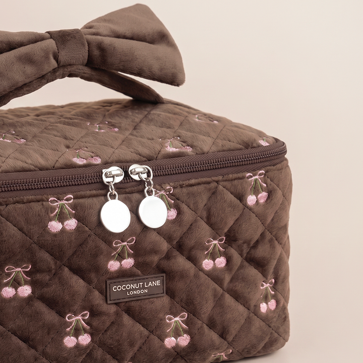 Close-up of a brown quilted velvet vanity case with pink embroidered cherries, a velvet bow handle and a “Coconut Lane London” label.