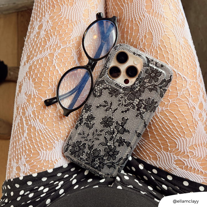 Clear Coconut Lane phone case with black lace design, placed beside eyeglasses on white lace tights and polka dot skirt.