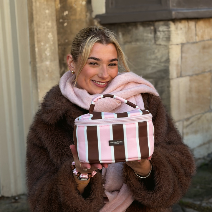 Woman with blonde hair, wearing a baby pink scarf and brown faux  fur jacket, holding a coconut Lane Cocoa & Pink Stripe round vanity makeup bag with chocolate brown and baby pink stripes.