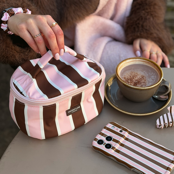 Woman wearing a baby pink scarf and brown faux fur jacket, holding a coconut Lane Cocoa & Pink Stripe round vanity makeup bag with chocolate brown and baby pink stripes. On the table beside this is the matching wavy phone case, claw clip and a mug of hot chocolate.