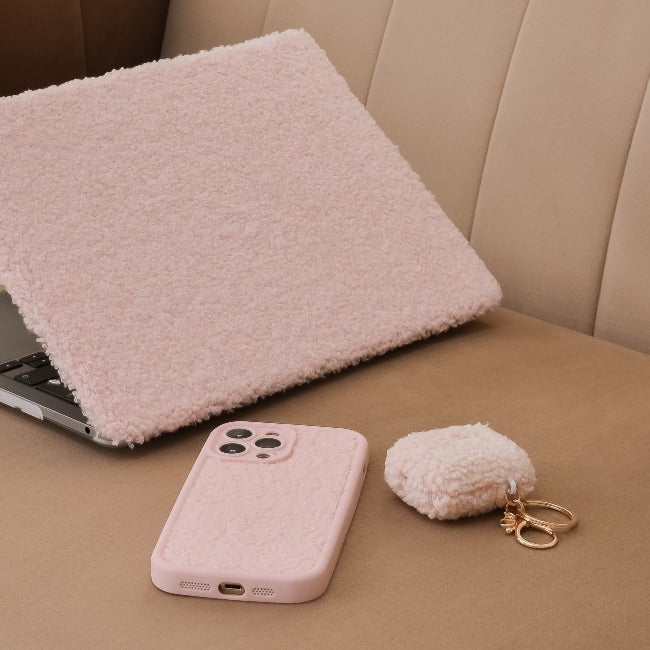 A pink fluffy laptop cover partially open next to a matching Cosy Teddy Phone Case - Pink by Coconut Lane, and a small fluffy keychain pouch with gold hardware. They are all placed on a beige cushioned bench, providing extra protective charm with their textured appeal.