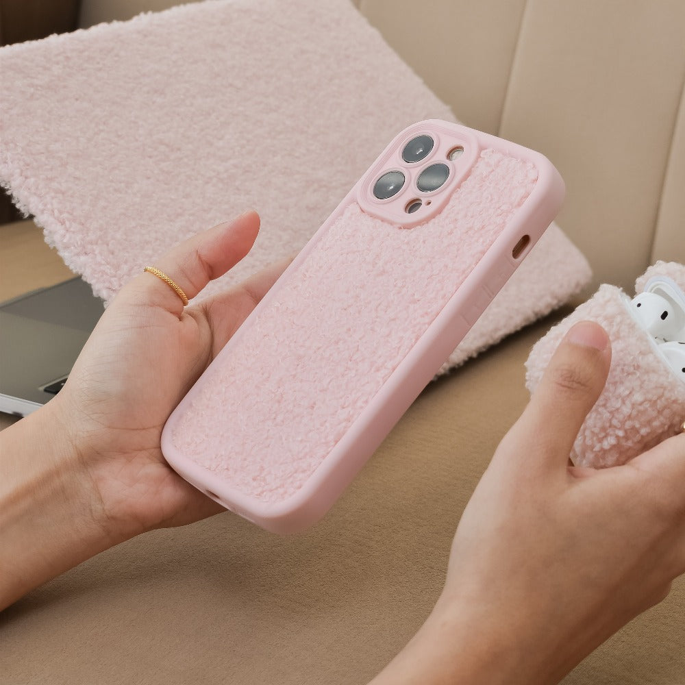 A person holds a Coconut Lane Cosy Teddy Phone Case - Pink, which has a soft, fabric-like texture. In the background, a matching pink tablet case and an extra protective pink AirPods case are visible on a beige surface. One of the person's hands also holds the pink textured AirPods case.