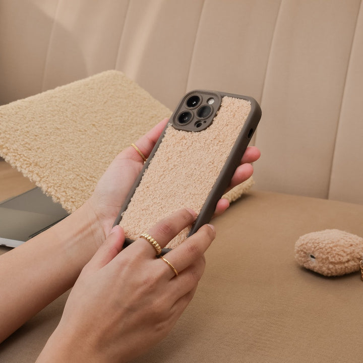 A person holding a Coconut Lane Cosy Teddy Phone Case in Salted Caramel is sitting on a beige sofa. The phone case has a soft, fuzzy appearance. In the background, there is a matching textured laptop sleeve and a small, fuzzy accessory on the sofa.