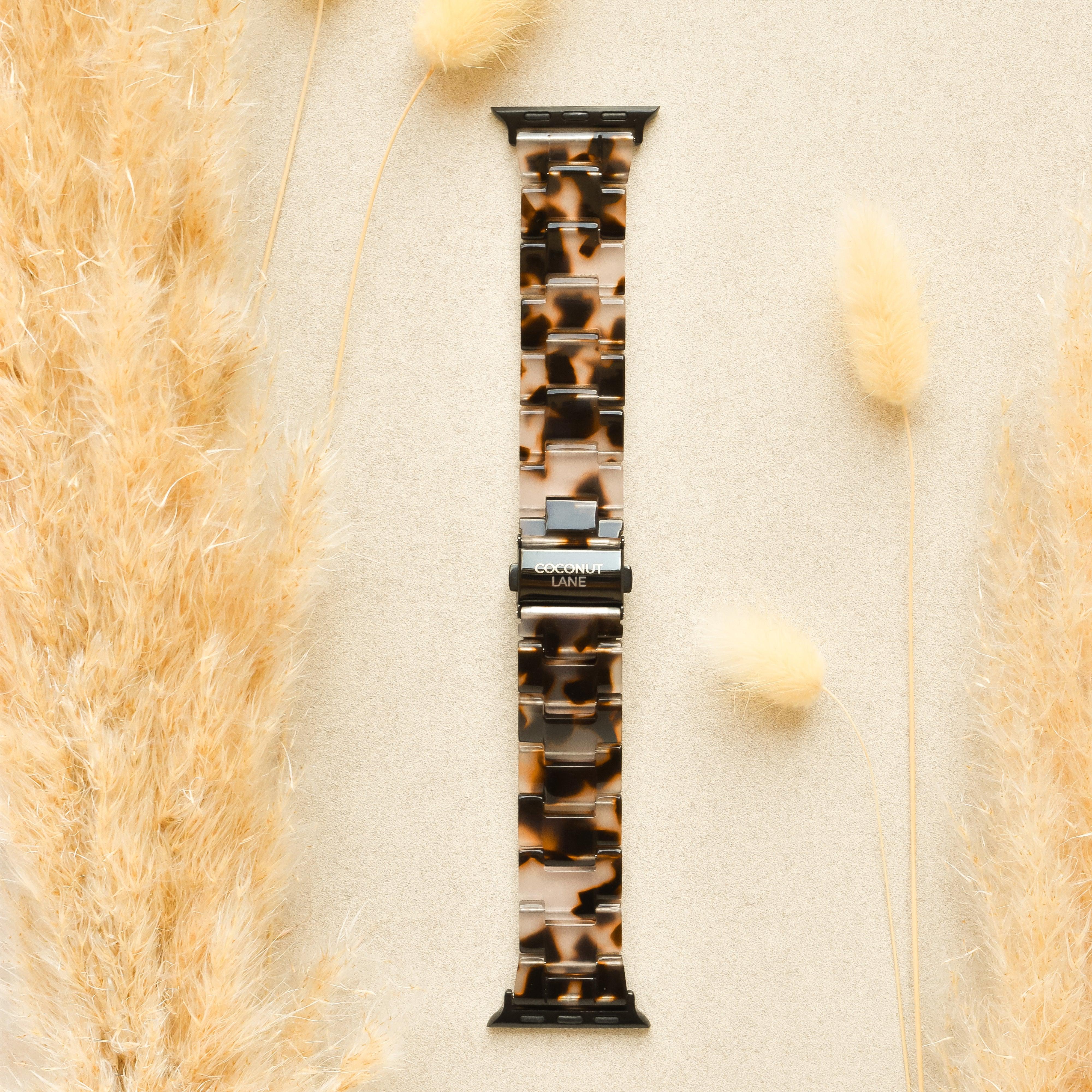 A Luxe Ivory Tort Apple Watch Strap from Coconut Lane is placed on a beige surface, surrounded by fluffy, beige ornamental grass on the left and right sides. The adjustable strap features silver connectors and prominently displays the brand name 