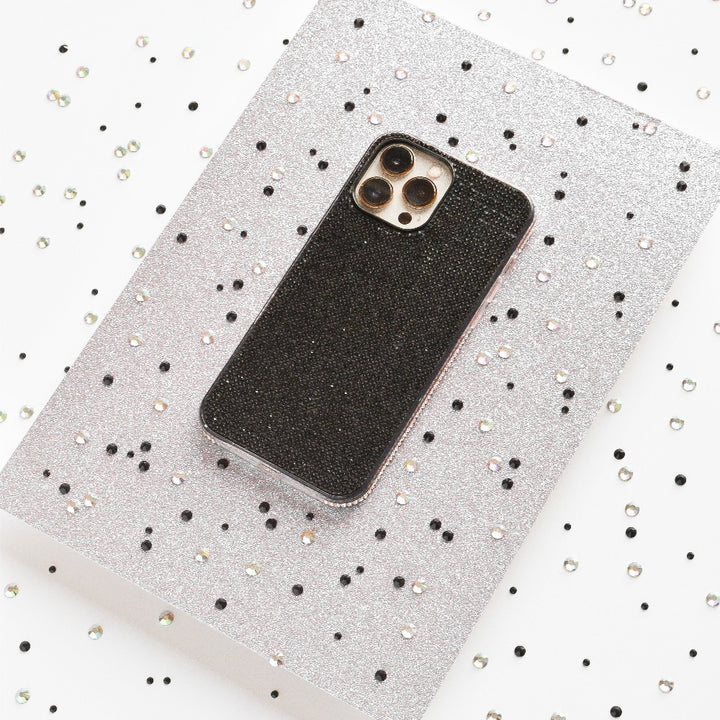 A smartphone with a Coconut Lane Disco Sparkle Phone Case - Black is placed on a silver glittery surface. The silver surface is adorned with scattered black and silver sequins, creating a festive and sparkly backdrop.