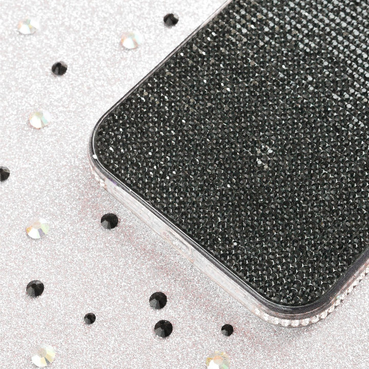 A close-up of a smartphone encased in Coconut Lane’s Disco Sparkle Phone Case - Black, resting on a silver, glittery surface. Both the extra protective case and the surface are adorned with scattered round, shimmering rhinestones.