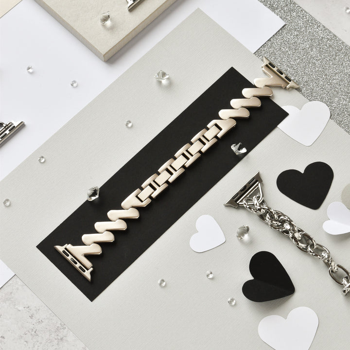 The Get Wavy Apple Watch Strap - Starlight from Coconut Lane is elegantly displayed on a black and white background, surrounded by heart-shaped cutouts and small decorative gems. Additional stainless steel metal adjustable links and clasps, suitable for all Apple Watch models, are creatively positioned around the central band.
