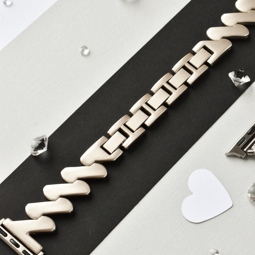 A close-up shot of the Get Wavy Apple Watch Strap - Starlight by Coconut Lane, laid diagonally on a black and white striped surface, surrounded by small, clear gemstones and a white heart cutout. The stainless steel adjustable watch band's interlocking links feature a matte finish. This strap is compatible with ALL Apple Watch models.
