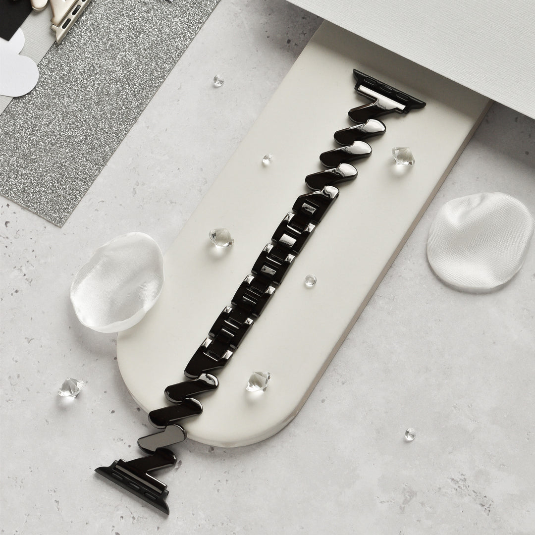The Get Wavy Apple Watch Strap - Black by Coconut Lane is elegantly showcased on a white rectangular holder. The holder is adorned with decorative silver glitter paper, white silk petals, and small clear beads, creating a sophisticated and refined display.