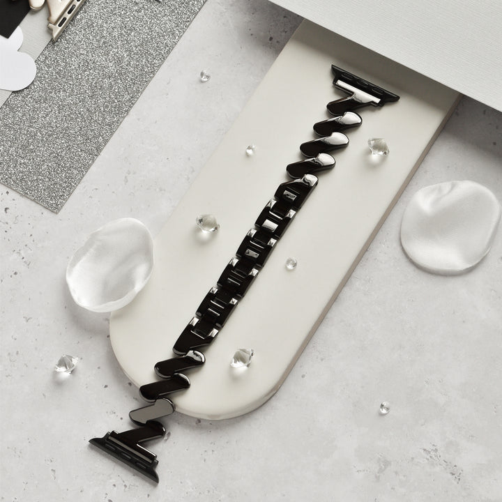 The Get Wavy Apple Watch Strap - Black by Coconut Lane is elegantly showcased on a white rectangular holder. The holder is adorned with decorative silver glitter paper, white silk petals, and small clear beads, creating a sophisticated and refined display.