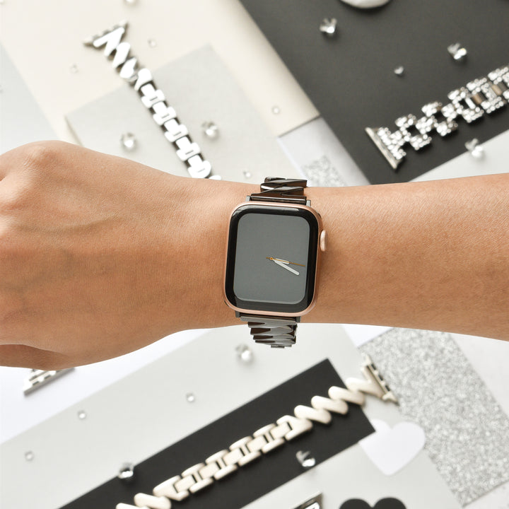 A person wearing a stylish Coconut Lane Get Wavy Apple Watch Strap - Black with an adjustable stainless steel strap. The smartwatch screen is off, reflecting light. The background features a collage of various geometric and textured elements in black, white, and silver.