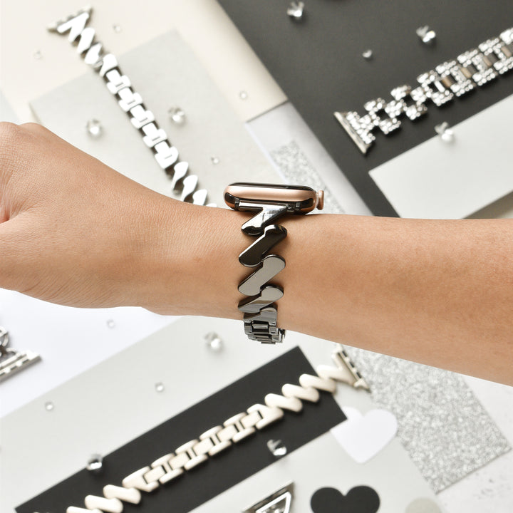 A hand wearing the Get Wavy Apple Watch Strap in black from Coconut Lane, featuring a stylish metallic, zigzag-patterned stainless steel band. Various other watch bands with different designs are displayed in the background on a flat surface, against a backdrop of black, white, and gray colors.