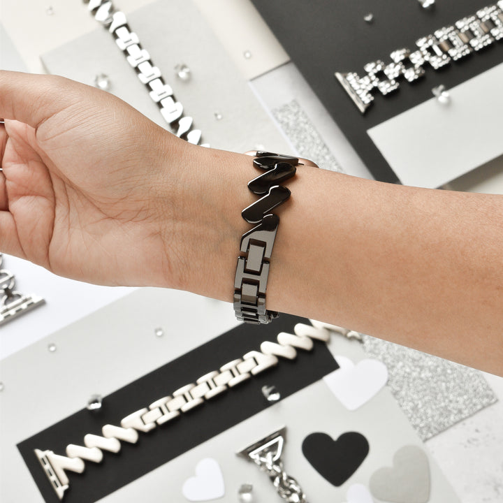 A person wearing the chic and robust Get Wavy Apple Watch Strap - Black by Coconut Lane on their wrist. The background showcases a variety of jewelry pieces, heart shapes, and sparkling decorations artistically arranged on different colored papers. The overall scene radiates a trendy and elegant vibe.