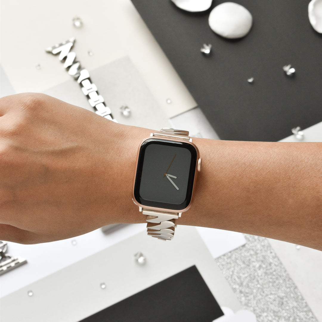 A close-up of a person’s wrist showcasing the sleek Get Wavy Apple Watch Strap - Starlight by Coconut Lane. The watch, which fits all Apple Watch models, has a black screen with a single white watch hand indicating the time. The background features a minimalist arrangement of geometric shapes in black, white, and silver.