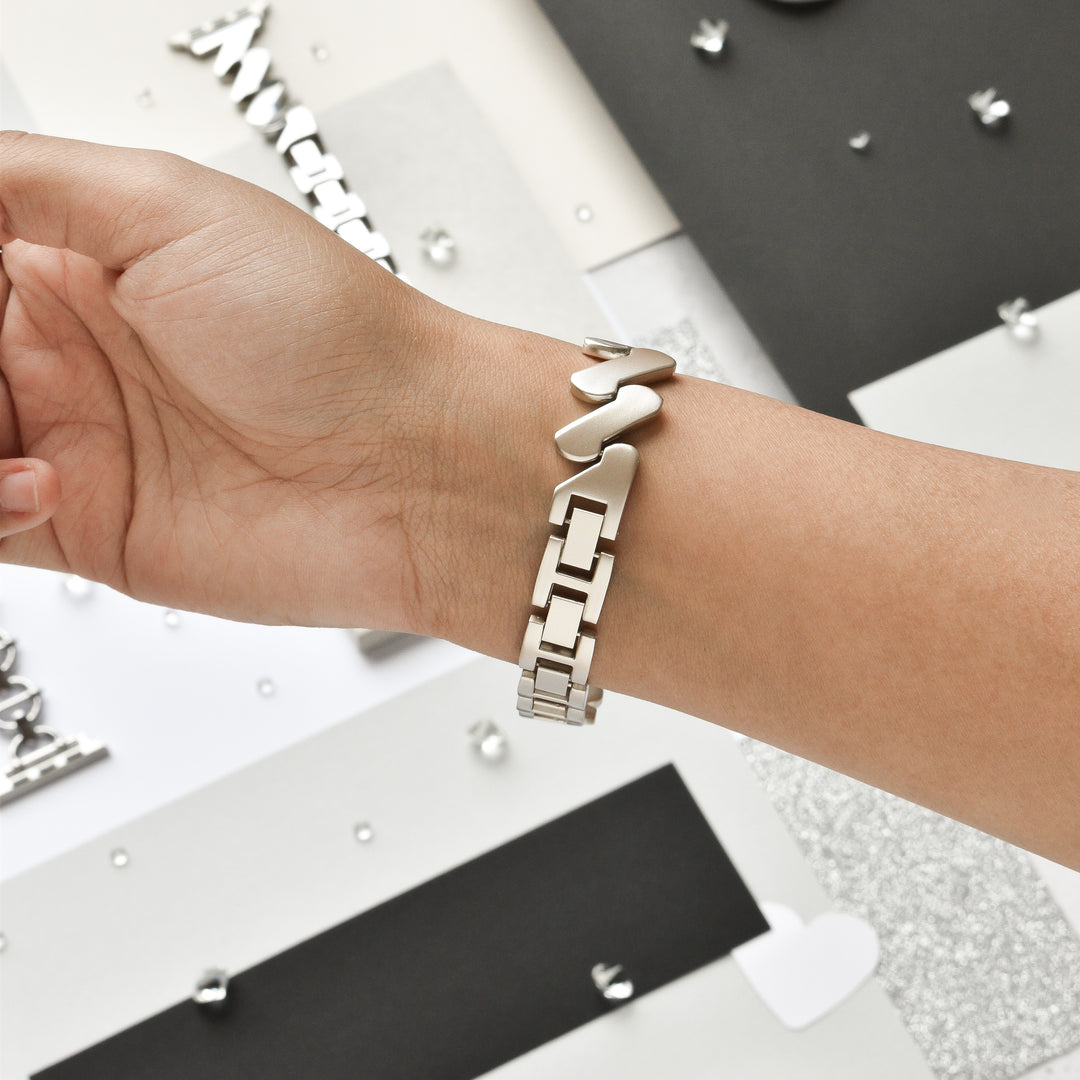 A person's wrist adorned with the Get Wavy Apple Watch Strap - Starlight by Coconut Lane, showcasing an elegant mix of heart-shaped and rectangular links. The backdrop features a flat lay of diverse geometric shapes and textures in black, white, and metallic hues. This Stainless Steel Metal Adjustable bracelet is compatible with all Apple Watch models.